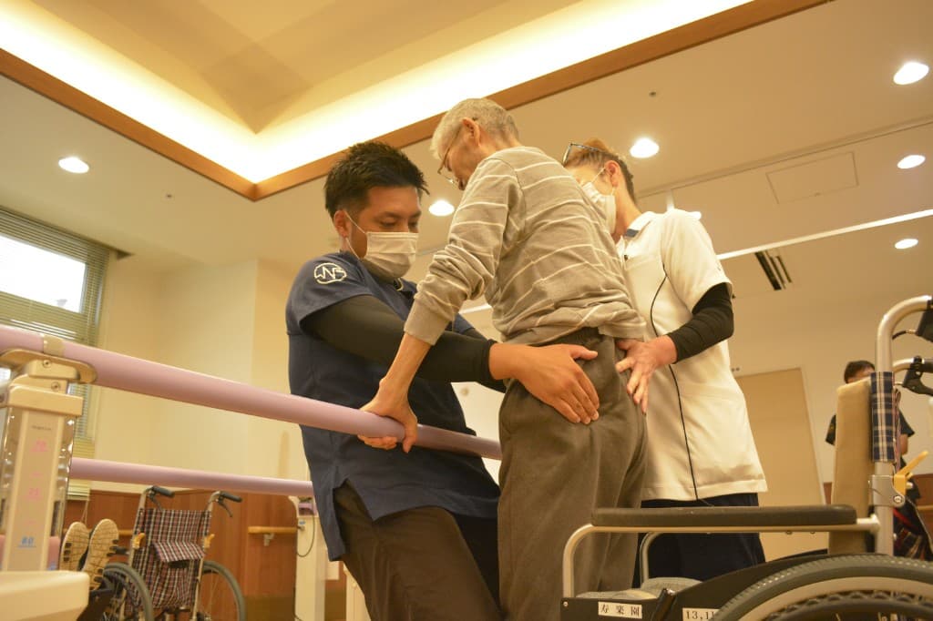 平行棒を使った立位・歩行訓練を、複数の専門職が支えている様子